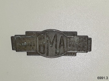 Badge is a rectangular shape, silver coloured metal, with embossed letters and basket-weave pattern