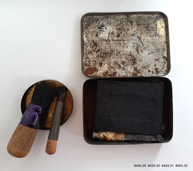 Two brushes, a reused tin lid for mixing, a block of ink and a tube of ink in a reused tin.
