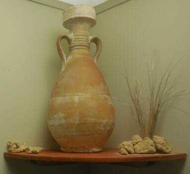 Amphora on a corner wooden shelf within the Mahogany Ship display, flanked by sandstone and grass. 