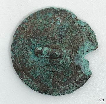 Metal disc, rough, irregular circle, raised area in centre of back containing small hole.