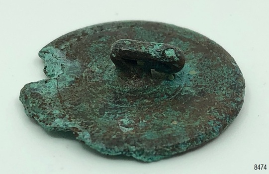 Metal disc, rough, irregular circle, raised area in centre of back containing small hole.