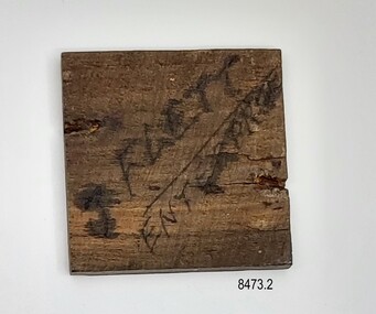 Small square of dark brown wood with handwritten inscription.
