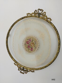 Round tray with a gilt brass border, filigree handles and embroidered doily encased in glass.