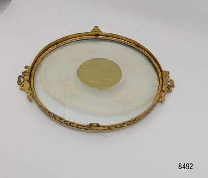 View from underneath of a round tray with a gilt brass border, filigree handles and embroidered doily encased in glass.