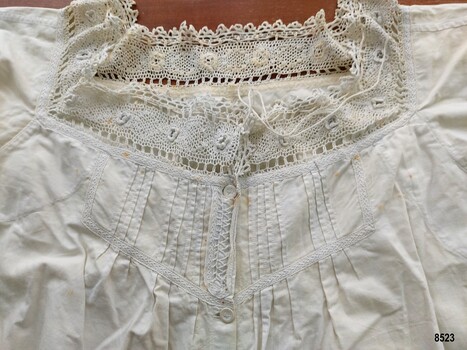 A square neckline with a drawstring cord, decorated with a crocheted lace trim in a floral design, pintucks and feather stitch embroidery.