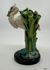 A white stork standing on one leg atop a  mound, an eel  in its beak, a frog beneath its foot, with water-lily pads.