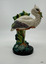 A white stork standing on one leg atop a  mound, an eel  in its beak, a frog beneath its foot, with water-lily pads.