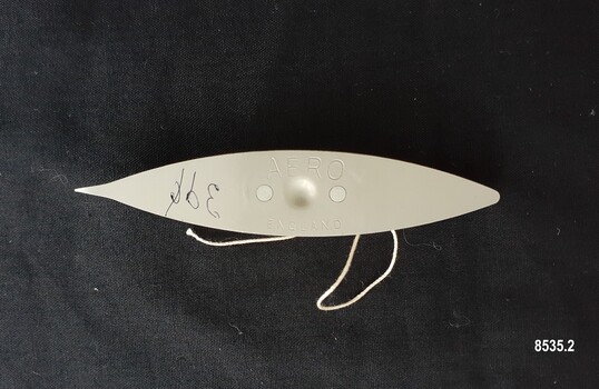 Beige, metal, boat shaped tatting shuttle with a small circular indentation in the centre and a length of thread emerging from the central bobbin