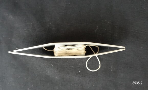 Side view of boat shaped, metal tatting shuttle showing the open ends and two internal posts that make up the bobbin (with thread attached)