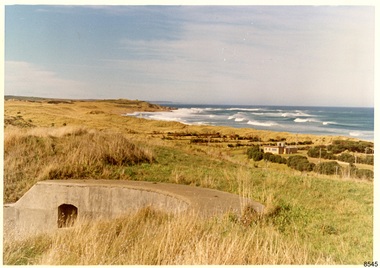 Curved concrete structure in grassy field, in front of tree-bordered fields, building, surf sea 