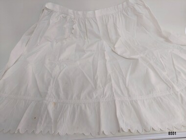 Short, tie waisted apron made of white cotton, featuring a scalloped trim on the frilled hem and pocket