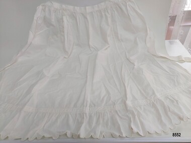 Long white apron with tie waist, scalloped and frilled hem and scalloped edge on pocket