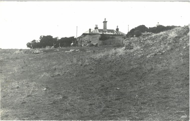 Black and white photograph of a fenced cottage with an iron roof. Nearby lighthouse on right.