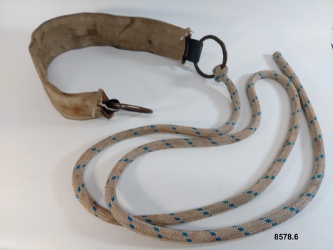 Wide leather strap covered with webbing. It has a metal ring at each end and a rope tied to one of the rings.