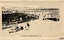 Beach with people in bathing costumes or protective clothing and hats, others in the shade.