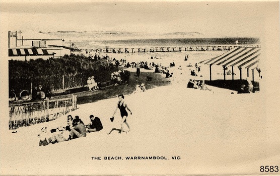 Beach with people in bathing costumes or protective clothing and hats, others in the shade.