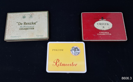 Three metal boxes, once containing cigarettes or cigars branded 'Ritmeester', 'Craven A' and 'De Reszke'.