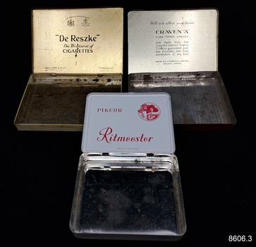 Three metal boxes, once containing cigarettes branded 'Ritmeester', 'Craven A' and 'De Reszke'.