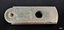 Silver cutter with measurements marked along one side a hole in the centre and the end, and inscriptions