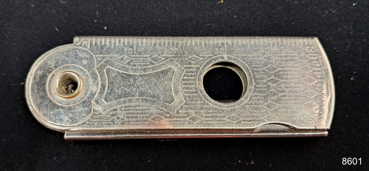 Silver cutter with measurements marked along one side a hole in the centre and the end, and inscriptions