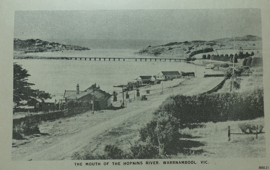 A road beside a river, buildings on the banks and a bridge between the banks.