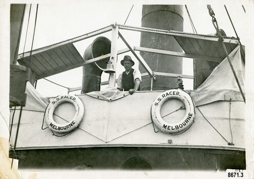 A man is leaning against the rail of a Tugboat. Two lifebuoys are tied to the side of the deck.