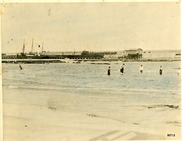 Colour tinted photograph of swimmers in front of a breakwater. A steamer is in the background. 