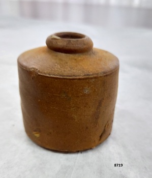 Brown clay inkpot with straight sides and small circular opening in the top. Some small dents and staining show on the surface of the sides.