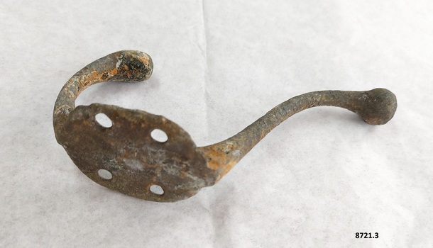 Metal coat hook with two prongs and four screw holes with a surface showing rust and corrosion.