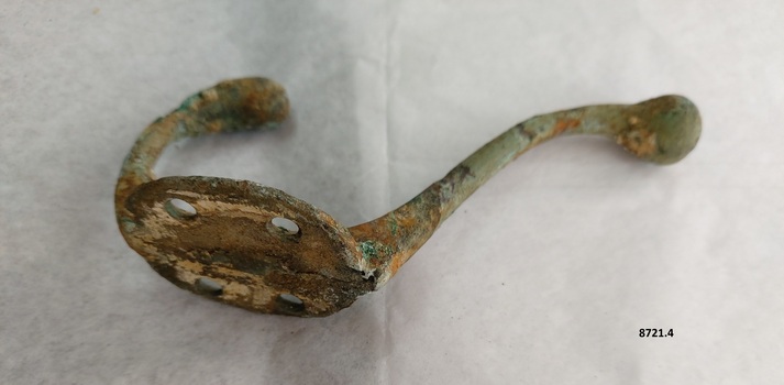 Brass coat hook with two prongs and four screw holes with a surface showing extensive green and brown oxidation.