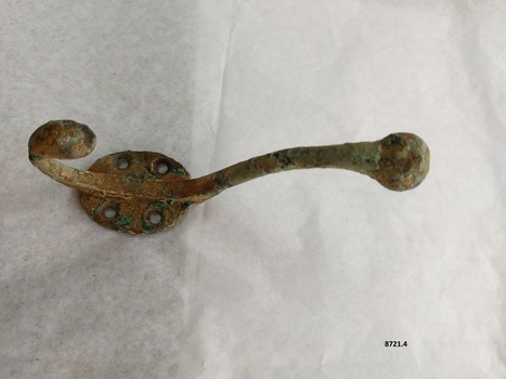 Brass coat hook with two prongs and four screw holes with a surface showing extensive green and brown oxidation.