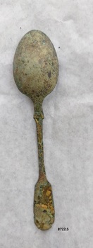 Back of dessert spoon with fiddle back handle. The thin metal has several small holes and is slightly encrusted with green and brown tarnish.
