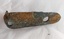 The outer side of an L shaped metal plate with one long, concave side (with two formed holes) and a narrow, short vertical arm with one hole. The outer side of the plate has a coating of green verdigris and a flaking layer of rust.