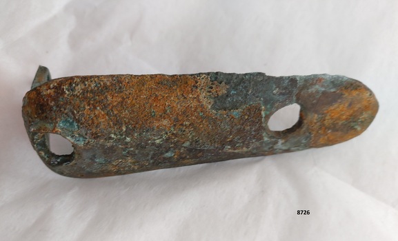 The outer side of an L shaped metal plate with one long, concave side (with two formed holes) and a narrow, short vertical arm with one hole. The outer side of the plate has a coating of green verdigris and a flaking layer of rust.