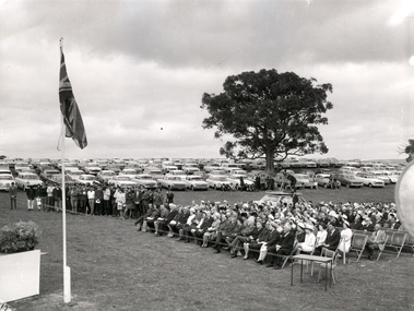 A seated audience, with cars in the background