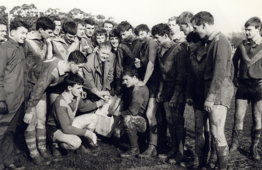 A football team and their coach poss for a photo on a muddy football ground.
