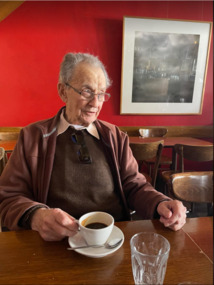 An elderly many sits at a table drinking coffee.