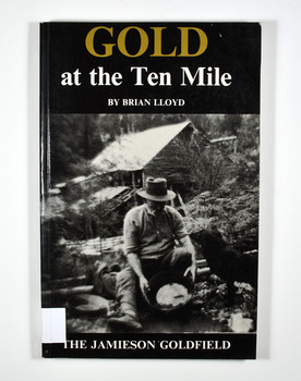 Paperback. Black cover. Front cover has a photograph of a man gold panning with his dog in a river. In the background is an old wooden house.