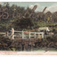 Shows a wooden, white painted bridge that crosses over a river. In the foreground, standing on the bank of the river, is a young girl wearing a hat. In the background is a timber building surrounded by wooden fences.  Birthday Greetings have been written in glitter across the image. On the reverse of the postcard is a message and an address handwritten in black ink. It appears to have had a postage stamp affixed but that has been removed.