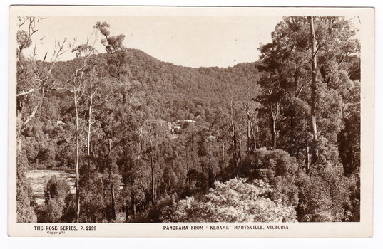 A black and white photograph of the view of Marysville from the "Kerami" guesthouse. Shows a glimpse of one or two buildings through the trees at the base of a heavily forested mountain in the background. In the foreground many trees. On the reverse of the postcard is a space to write a message and an address and to place a postage stamp. The postcard is unused.