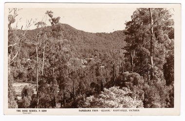 A black and white photograph of the view of Marysville from the "Kerami" guesthouse. Shows a glimpse of one or two buildings through the trees at the base of a heavily forested mountain in the background. In the foreground many trees. On the reverse of the postcard is a space to write a message and an address and to place a postage stamp. The postcard is unused.