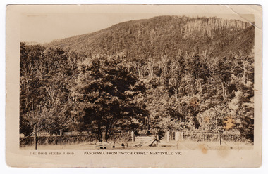 Shows a panoramic view from Wych Cross which was a guesthouse in Marysville. In the foreground is a large tree with a wooden bench seat at the base. Behind the tree can be seen the entrance gate and fenceline of the property. In the background there is a heavily forested mountain. On the reverse of the postcard is a handwritten message.