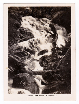 Shows the Cora Lynn Falls near Marysville in Victoria. Shows the falls cascading down the mountain surrounded by several boulders. There is a fallen log across the falls. The title of the photograph is along the lower edge of the photograph.