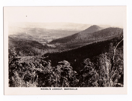 Shows the view of the surrounding hills from Nichol's Lookout which is on the Marysville-Wood's Point road near Marysville in Victoria. In the background are heavily forested mountains. The title of the photograph is along the lower edge of the photograph.