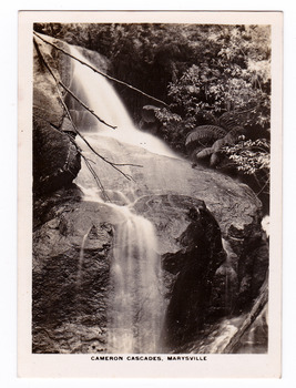 Shows the Cameron Cascades  near Marysville in Victoria. Shows the waterfall cascading down over some large boulders. In the background can be seen trees and tree ferns. The title of the photograph is along the lower edge of the photograph.
