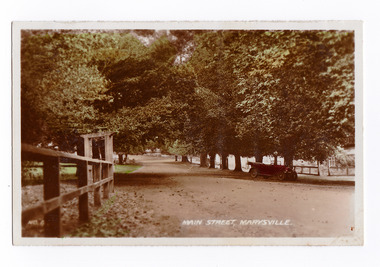 Shows the tree lined main street in Marysville. In the foreground on the left of the photograph can be seen some wooden fencing with an entrance lynch gate. In the right of the photograph can be seen an early model red car standing under a tree. The title of the photograph is handwritten in white ink along the lower edge.