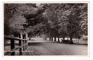  Shows the tree lined main street in Marysville. In the foreground on the left of the photograph can be seen some wooden fencing with an entrance lynch gate. In the right of the photograph can be seen an early model car standing under a tree. The title of the photograph is handwritten in white ink along the lower edge. On the reverse of the postcard is a handwritten message.
