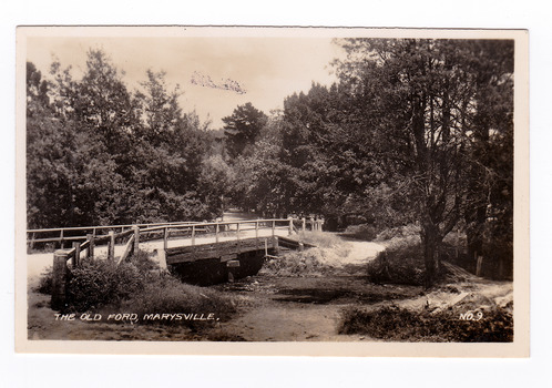 Shows the old ford in Marysville, Victoria. Shows a wooden bridge over the river. In the background the heavily treed road leads off into the distance. The title of the photograph is handwritten in white ink along the lower edge.