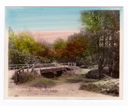 An early colour tinted photograph of the old ford in Marysville, Victoria. Shows a wooden bridge over the river. In the background the heavily treed road leads off into the distance.The title of the photograph is handwritten in white ink on the lower edge.