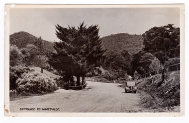 Shows the view looking up the Marysville-Wood's Point Road towards the corner with Murchison Street in Marysville in Victoria. Shows an unsealed road leading off to the left. There is a glimpse of another road leading off to the right. In the foreground there are shrubs and large trees on the left hand side of the road and on the right there is a parked car. In the background are heavily forested mountains. The title of the photograph is handwritten in black ink on the lower edge. On the reverse of the postcard is space to write a message and an address and to place a postage stamp. The postcard is unused.
